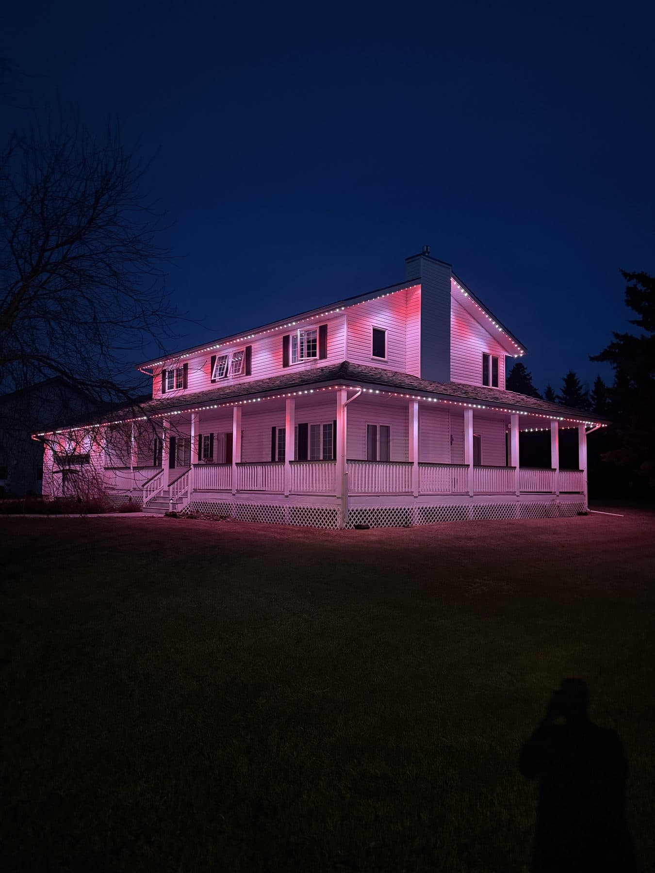 Permanent LED lighting on a large farmhouse with a wraparound porch at dusk